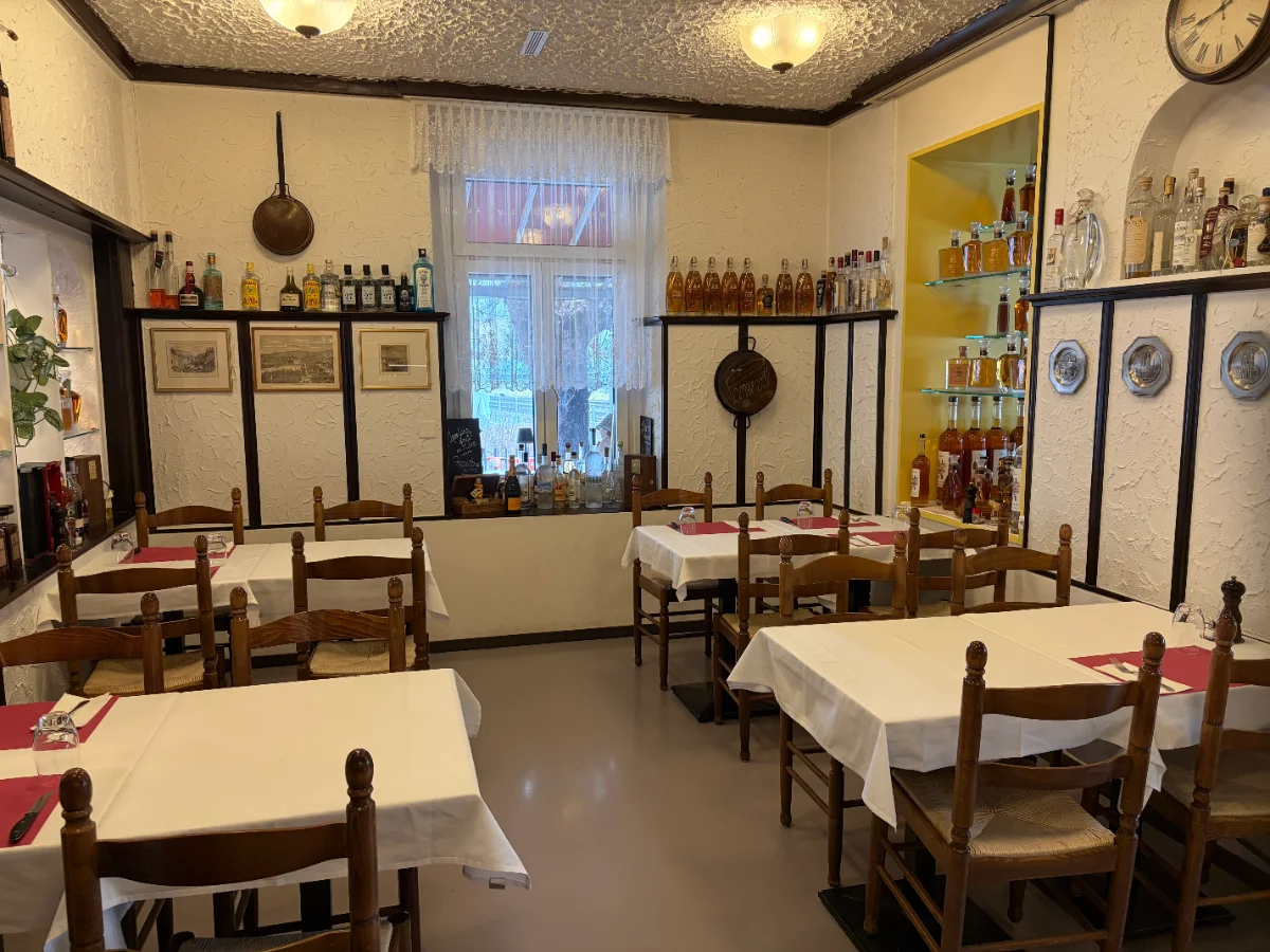 Innenansicht eines Restaurants mit Tischen, Stühlen und dekorativen Weinflaschen an der Wand.